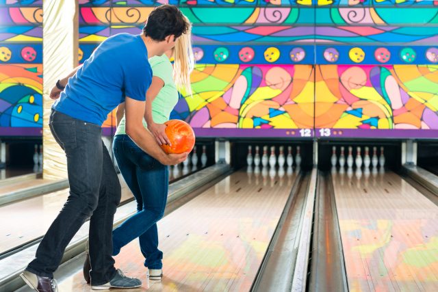 Young couple or friends, man and woman, playing bowling with a ball in front of the ten pin alley, he shows her how it works